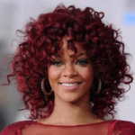 Person with curly red hair, wearing a red top, and hoop earrings, smiling at the camera.
